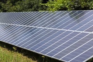 Close-up surface of lit by sun modern saving efficient stand -alone blue shiny solar photo voltaic panels system producing renewable clean energy on green grass and trees foliage background.